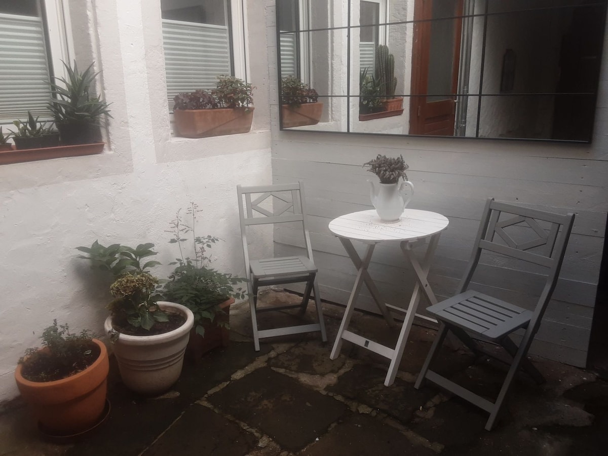 A small outdoor seating area features two gray chairs and a round white table. Potted plants are arranged around the space, adding greenery. Mirrors reflect the natural light, enhancing the bright ambiance of this intimate corner.