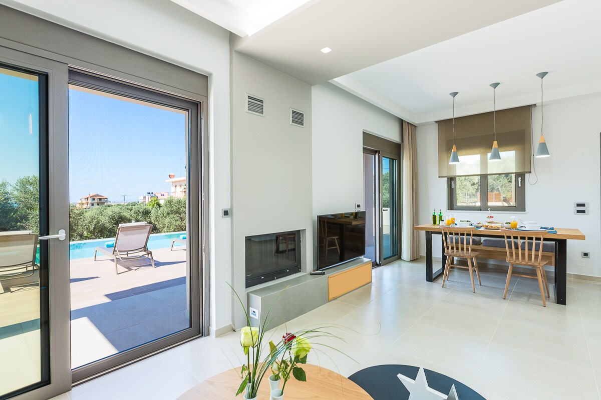 The open-plan living area features large glass doors that provide a view of the outdoor pool and garden. A modern dining table is set for meals, while a comfortable lounge area is positioned nearby. Natural light enhances the spaciousness of the room.