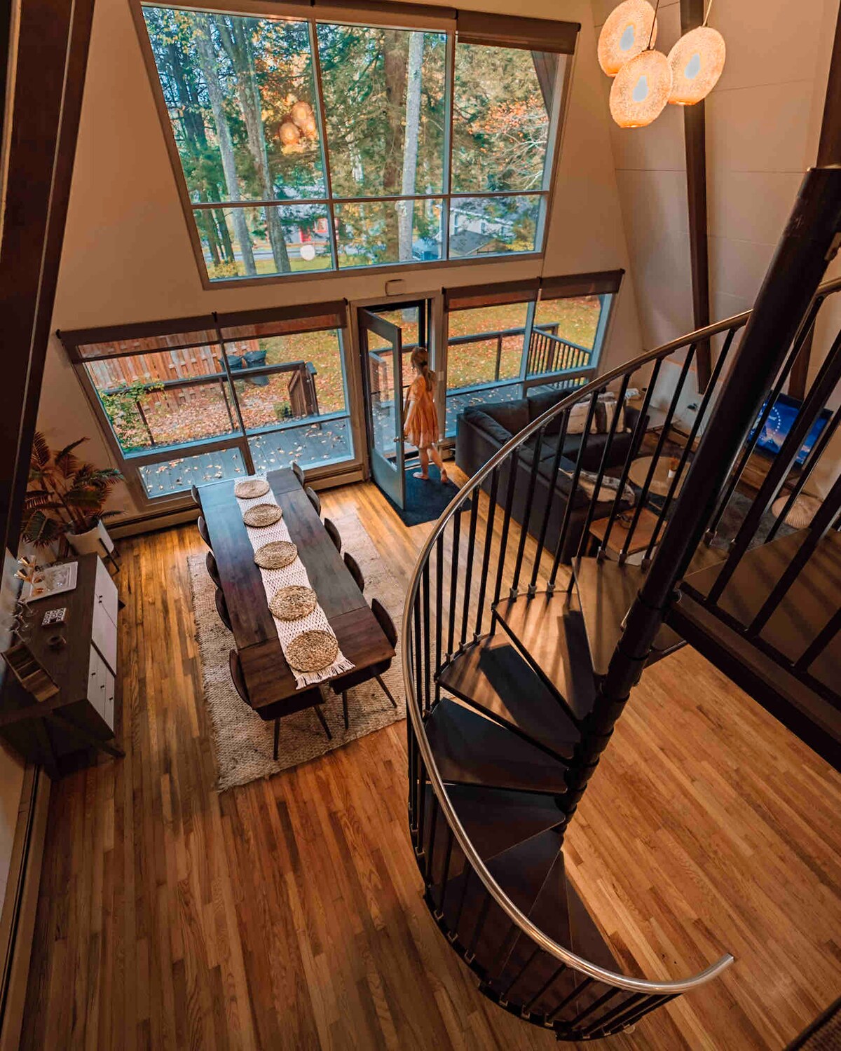 An aerial view showcases the open-concept main floor, featuring a dining table set for several guests and a cozy living area with a large window overlooking fall foliage. A spiral staircase leads to the upper floors, complemented by unique light fixtures and warm wood flooring.