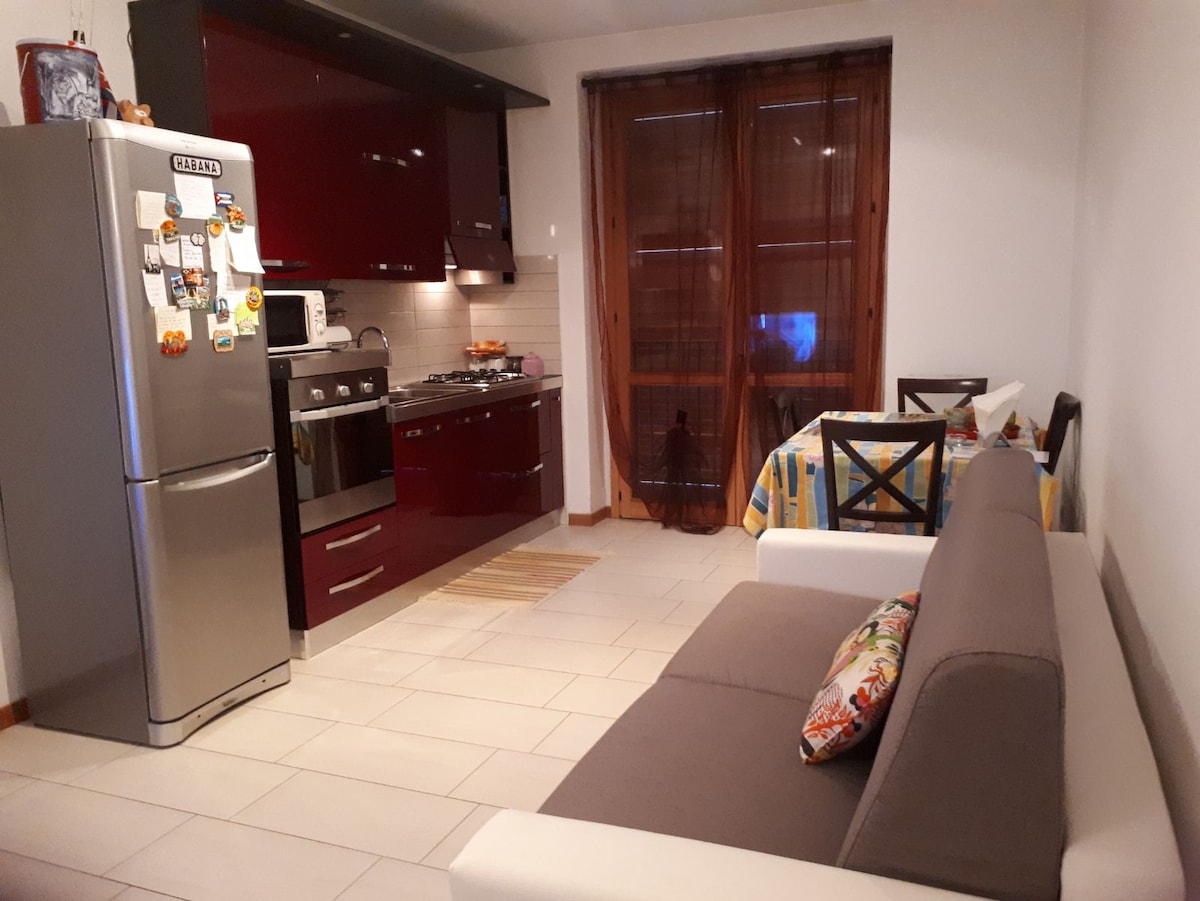 A modern kitchen area features sleek red cabinetry and stainless steel appliances. A spacious gray sofa sits next to a dining table with colorful tableware. Natural light filters through the brown curtains, illuminating the tiled floor.