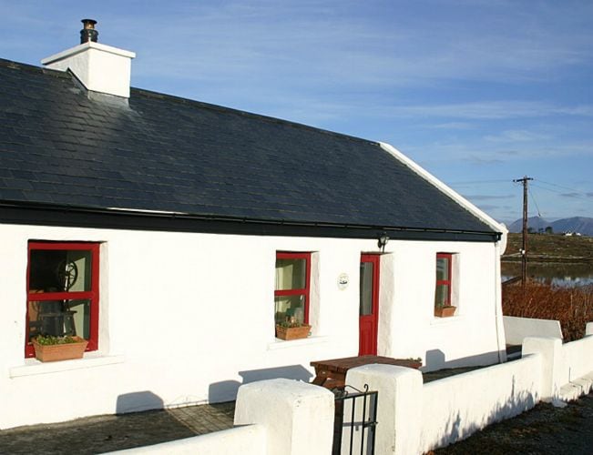 A traditional cottage showcases a white exterior with a slate roof. Windows with red frames provide a pop of color, while flower pots add charm. The entrance features a red door, and a low stone wall borders the property, enhancing the welcoming appearance.