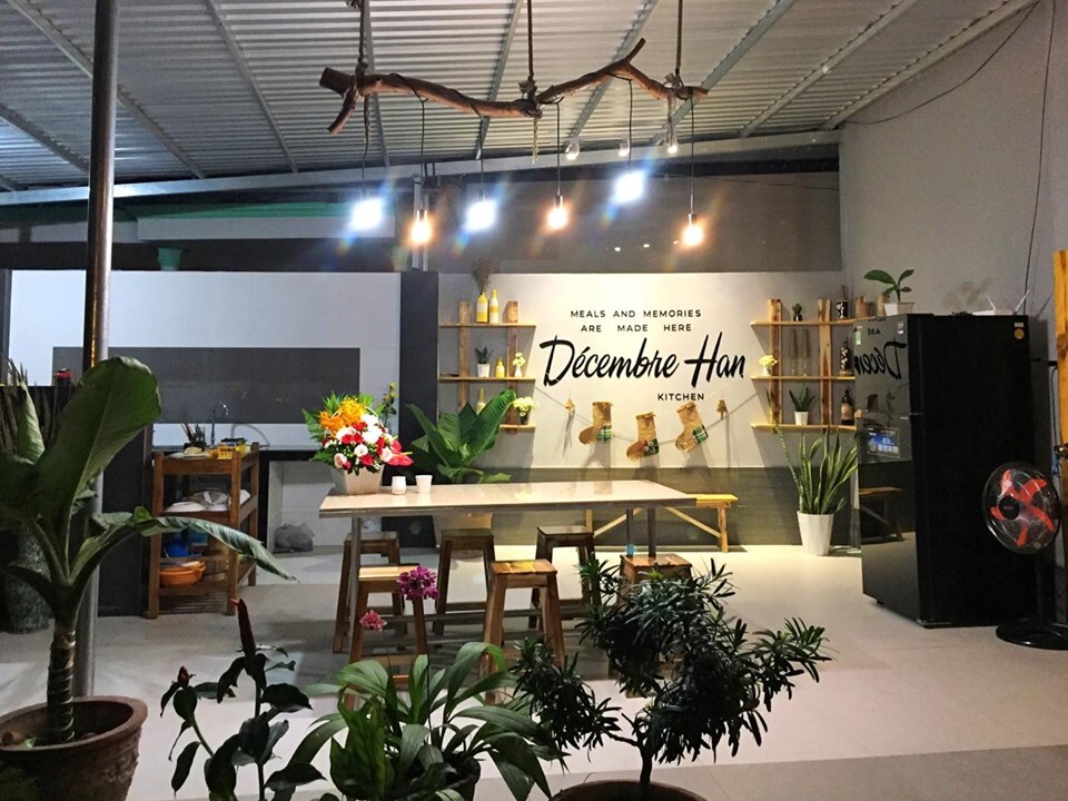 A spacious outdoor kitchen area is featured, highlighted by a central wooden dining table surrounded by plants. Decorative shelves display handmade items, and a wall adorned with the name 'Décembre Han' adds a personal touch. Ambient lighting enhances the inviting atmosphere.