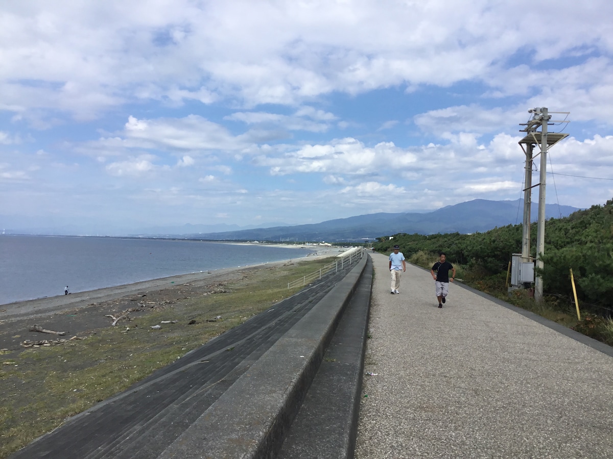 A scenic pathway is visible along the coastline, bordered by gentle waves on one side and greenery on the other. The horizon stretches towards distant mountains under a sky filled with scattered clouds. Two individuals can be seen walking along the path.