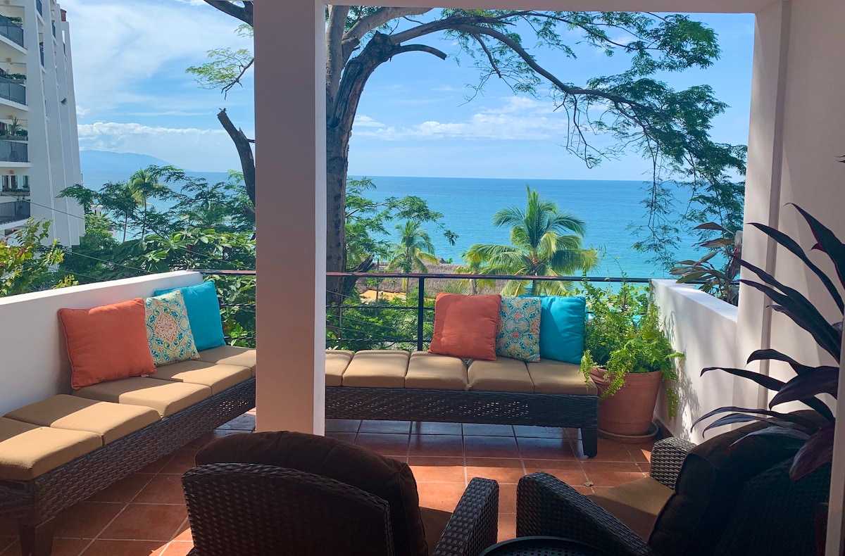 A private outdoor terrace features a comfortable seating area with plush cushions, offering views of the ocean and surrounding greenery. The vibrant colors of the pillows contrast with the neutral tones of the furniture, all framed by a clear sky above.
