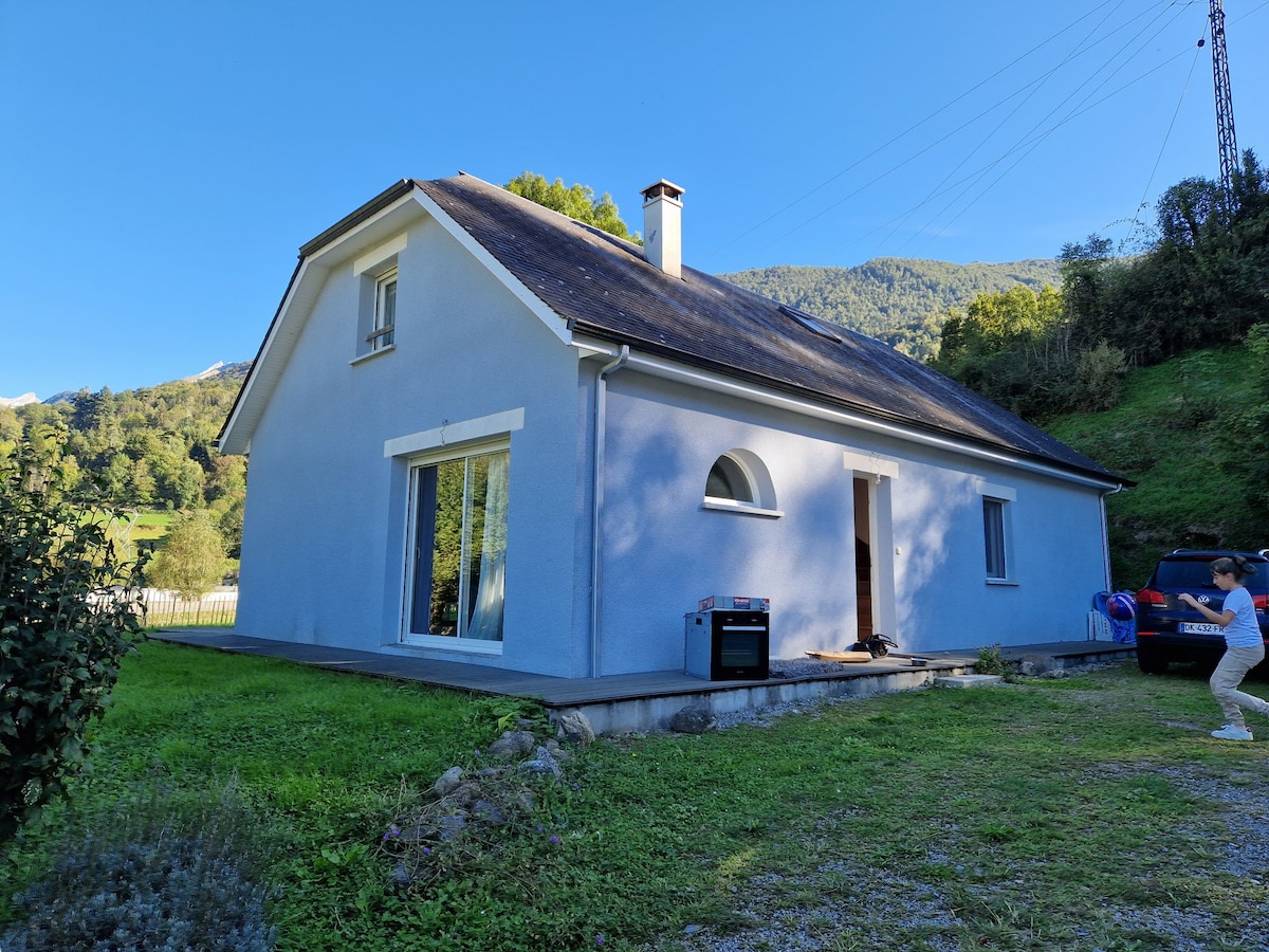 A single-story house is set against a backdrop of mountains and greenery. The exterior features pale blue walls and large windows, providing natural light. The property has a small outdoor area with grass and gravel, suitable for parking vehicles.
