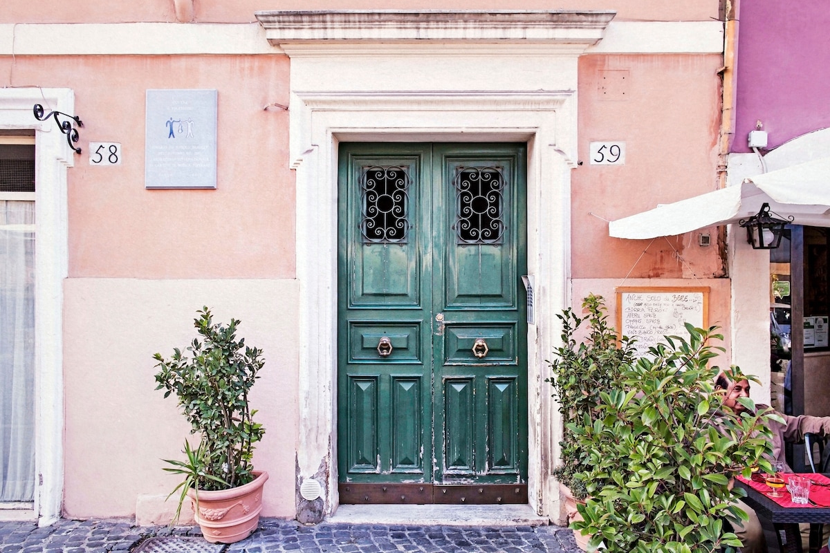 The entrance features a pair of green doors framed by a light-colored facade. Intricate details adorn the doors, complemented by decorative wrought iron elements. Potted greenery flanks the entrance, enhancing the inviting appearance of this historic building in Trastevere.