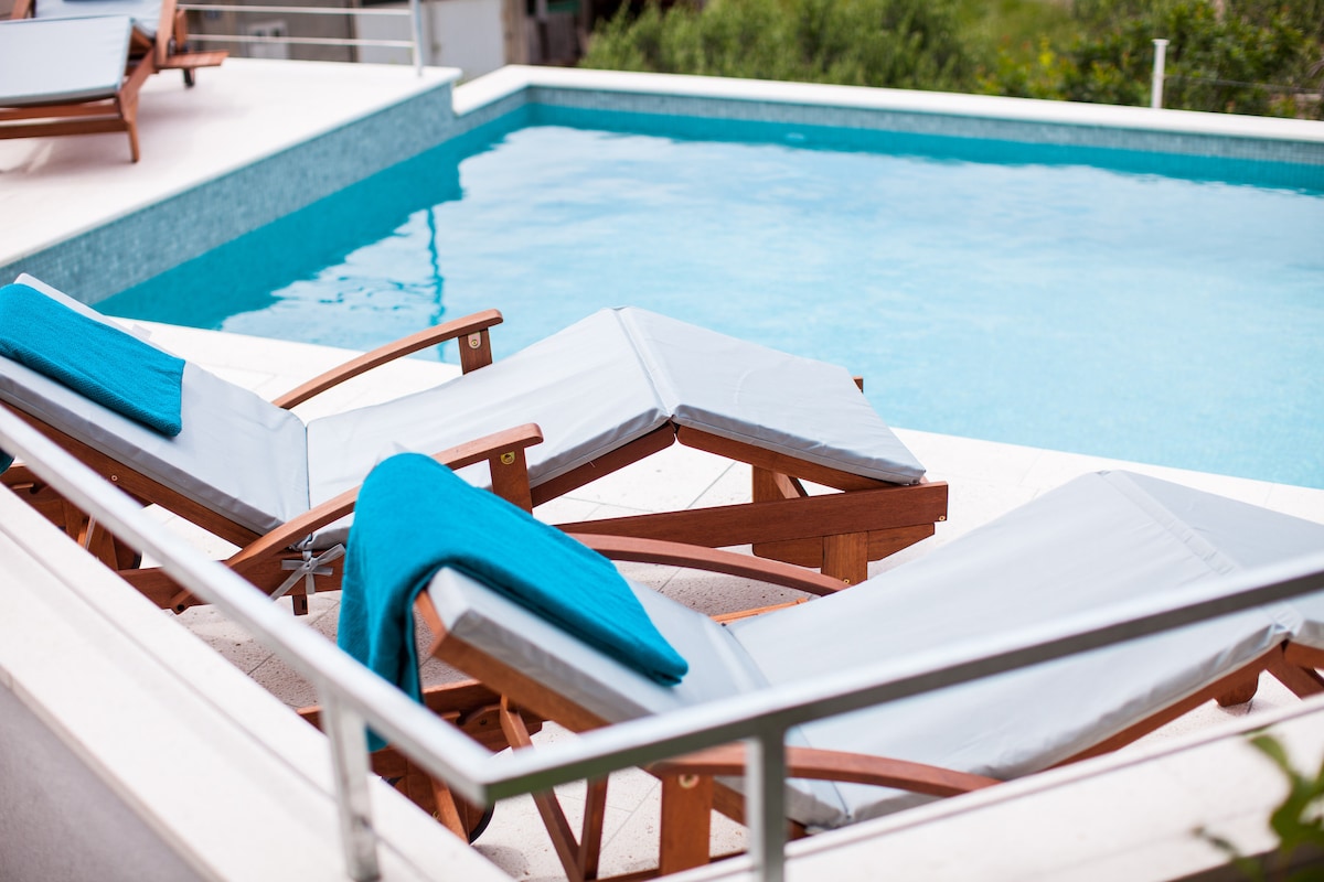 Two sun loungers with light gray cushions and blue towels are positioned beside a clear swimming pool. The pool has a smooth tiled edge, reflecting the greenery of the surrounding area. The relaxing environment is enhanced by a spacious sun deck.