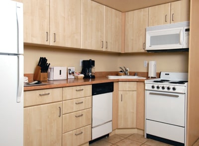 A functional kitchen area is equipped with a refrigerator, stove, and microwave. Light wood cabinetry creates a bright environment while counter space accommodates a coffee maker and utensils. The tile flooring adds an easy-to-clean surface, enhancing the kitchen's practicality.