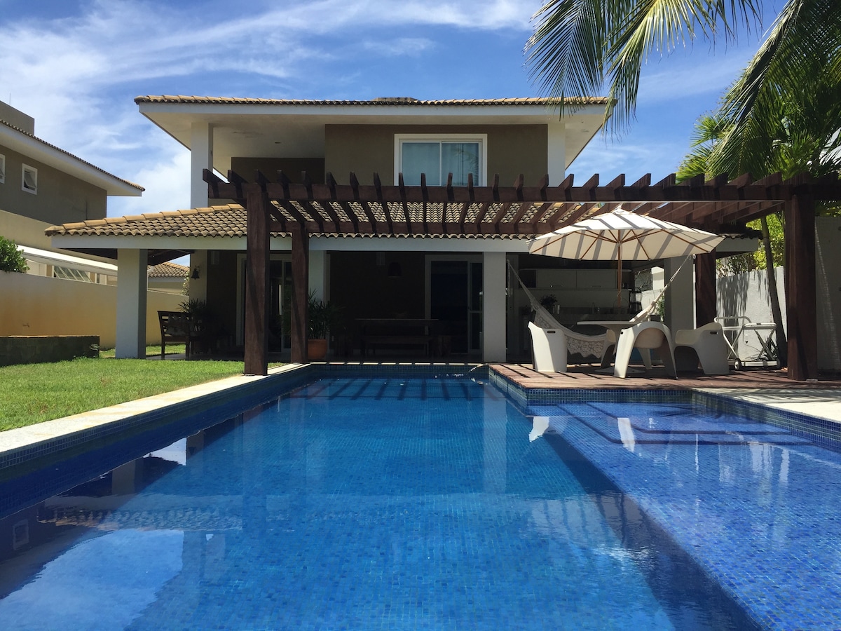 A spacious outdoor area is highlighted by a clear blue swimming pool. Comfortable seating arrangements are situated under a shaded pergola, with a large umbrella providing additional cover. Lush green grass and palm trees frame the setting, complementing the house's architecture in the background.