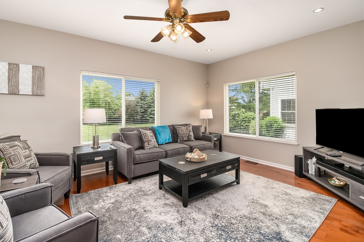 A comfortable living room is presented with a sectional sofa, armchairs, and a coffee table. Natural light streams through large windows, illuminating the space. A light-colored area rug covers the hardwood floor, while a television and decorative items are positioned on a media console.