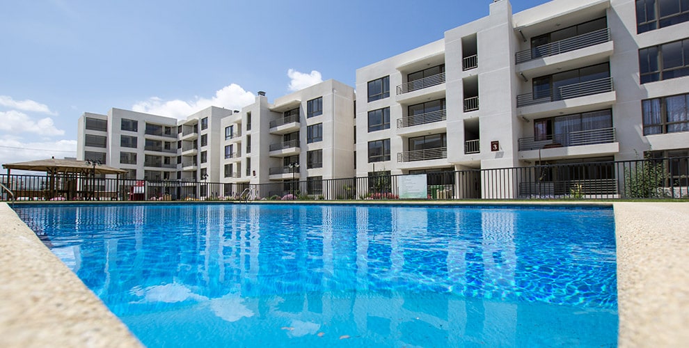 The image captures a clear blue swimming pool in the foreground, reflecting the sunny sky. In the background, several modern apartment buildings with balconies are visible, framed by well-maintained greenery and a fenced perimeter.