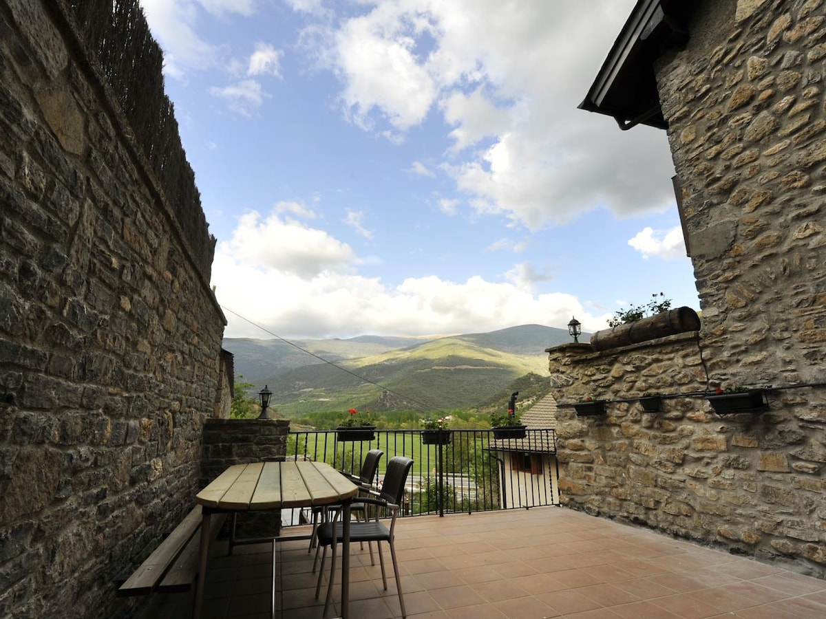 A spacious terrace features a large wooden table surrounded by chairs, offering views of the mountainous landscape. The stone walls provide a rustic charm, while the sky above displays varying shades of blue and soft clouds, enhancing the overall outdoor experience.