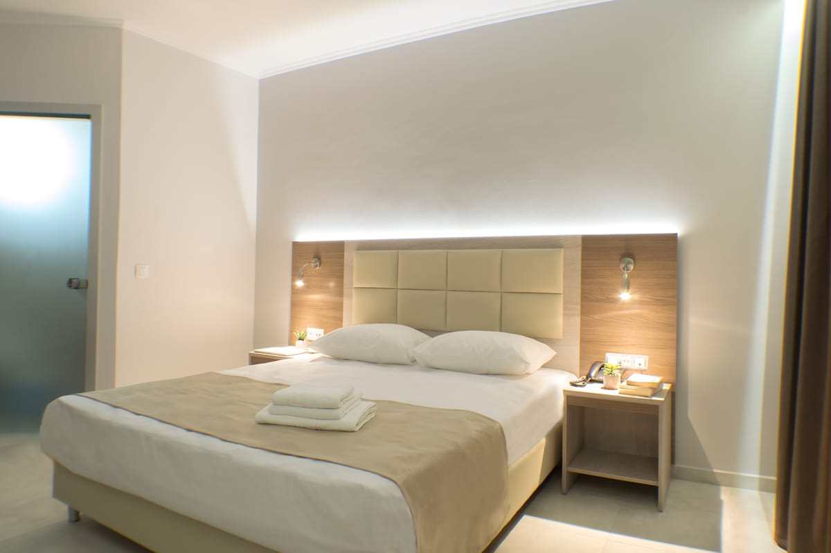 A spacious sleeping area features a light-colored bed with a beige blanket and neatly arranged white towels. Soft lighting is provided by wall-mounted fixtures on either side of the headboard, creating a calm atmosphere. Light-colored walls complement the light tile flooring.