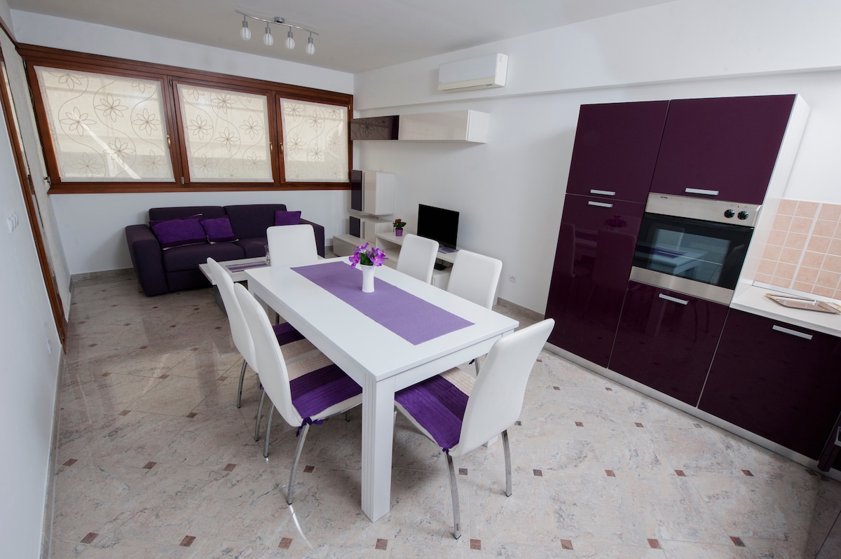 A modern living space is presented with a purple-accented theme. A dining table with white chairs is centered, complemented by a purple table runner. A dark purple sofa is visible in the corner, with large windows allowing natural light to enhance the room.