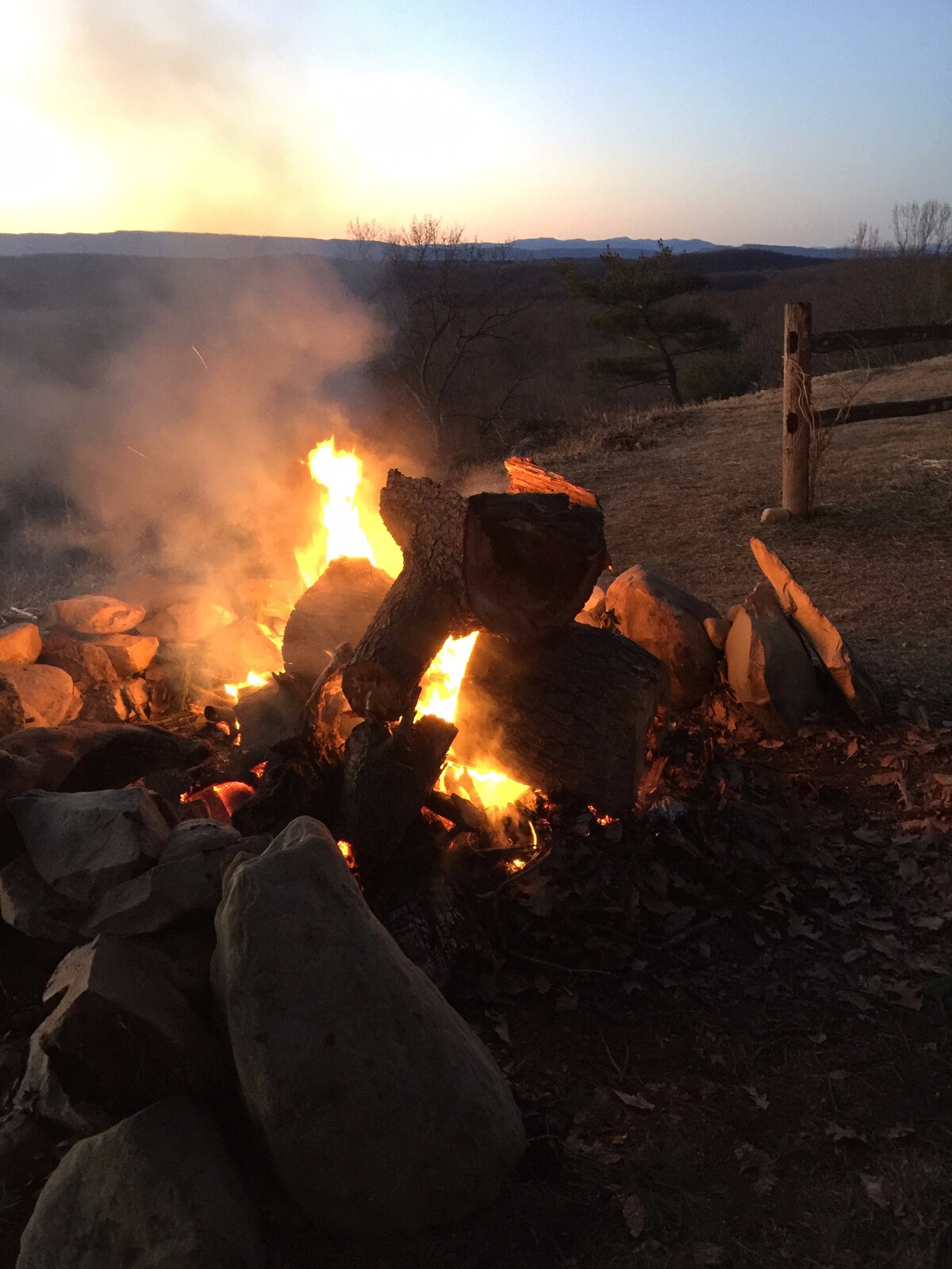 A campfire is seen surrounded by large stones, with flames dancing upward against a twilight sky. Smoke rises gently, adding to the atmosphere. In the background, silhouettes of distant mountains can be spotted as the sun sets.