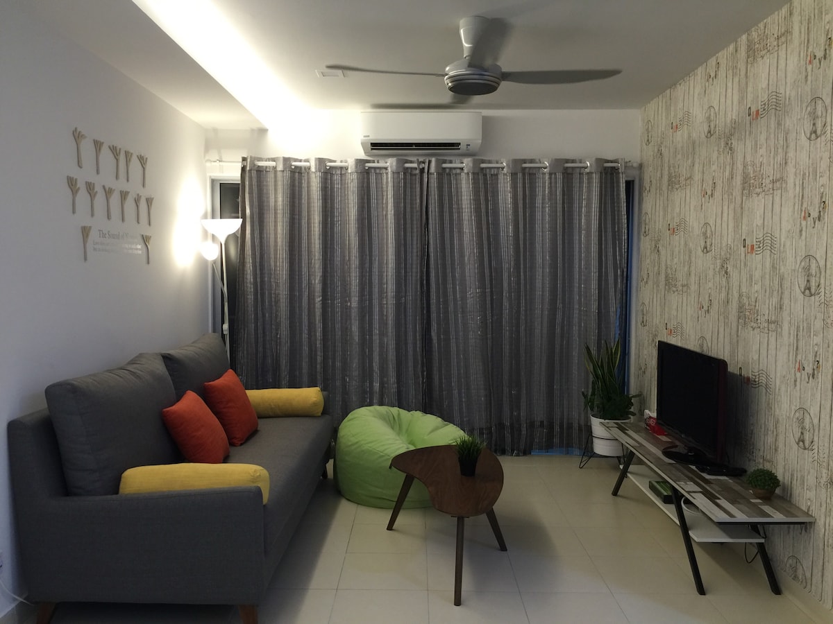 A modern living area features a gray sofa adorned with orange and yellow cushions. A small wooden coffee table sits in front of a low TV stand, while a green bean bag adds a playful touch. Large windows are dressed with sheer gray curtains, allowing soft light in.