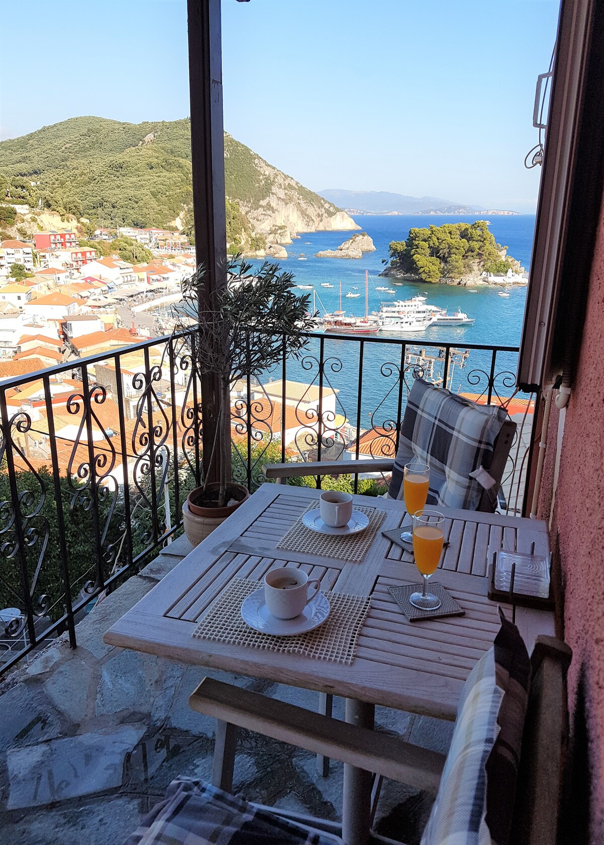 A balcony view showcases a charming breakfast setup with two glasses of orange juice and a cup of coffee on a wooden table. The backdrop features Parga's colorful town and the scenic coastline, offering panoramic views of the harbor and surrounding greenery.