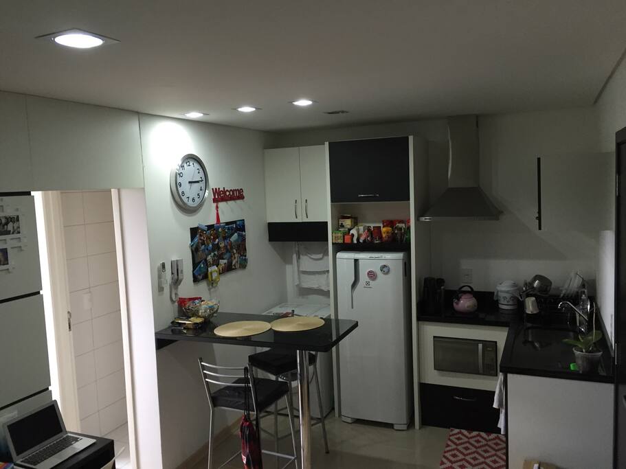 Foto da sala e cozinha onde tem fogão, microondas e todos utensílios de cozinha. Taça de vinhos também. Máquina de lavar e secar roupas.