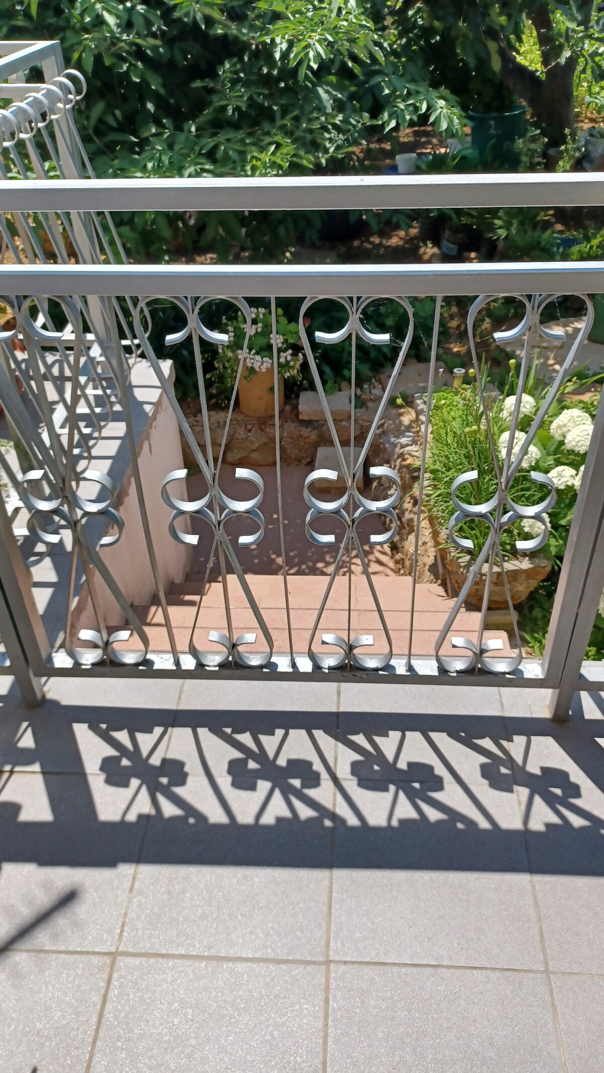 A decorative metal railing features intricate scrollwork, casting unique shadows on the tiled surface below. The background reveals a garden with lush greenery, adding a vibrant touch to the view from the balcony. Natural light enhances the details of the railing and surroundings.