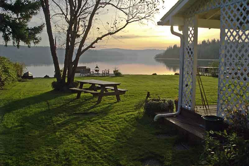 A serene lakefront view is captured at dawn, showcasing a lush green lawn bordered by trees. A wooden picnic table sits on the grass, while a private dock extends into the calm waters, reflecting the soft morning light.