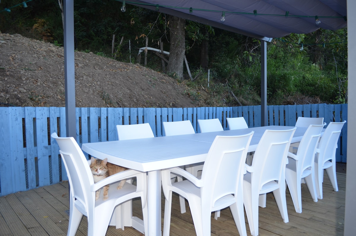 An outdoor dining area is furnished with a large rectangular table surrounded by multiple white plastic chairs. A wooden deck is visible beneath the furniture, and a blue wooden fence provides a backdrop. Lush greenery is present in the surrounding landscape.