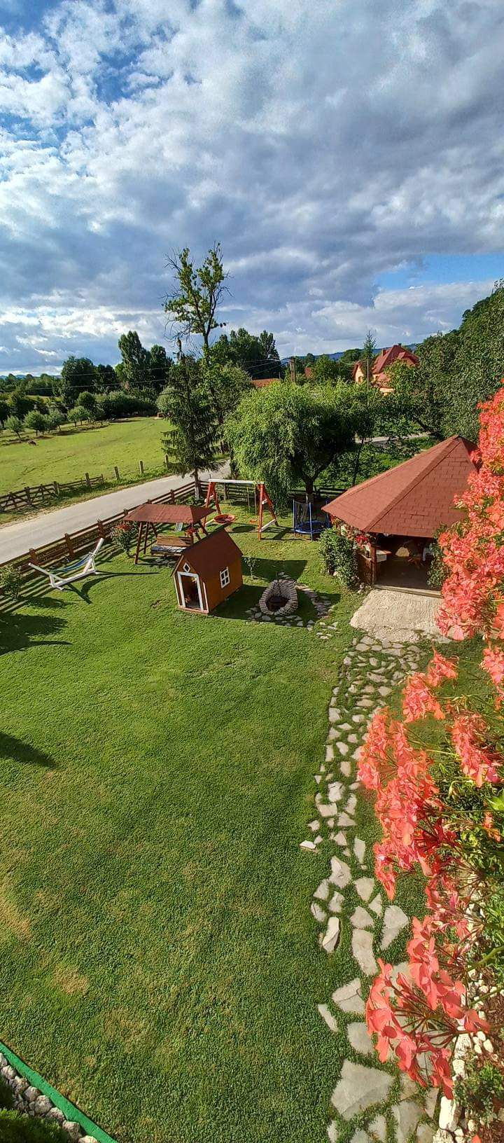A lush green yard features a charming play structure and a small wooden house surrounded by vibrant flowers. The landscape is dotted with trees and offers a view of the countryside, while a stone pathway leads to a cozy wooden building with a sloped roof.