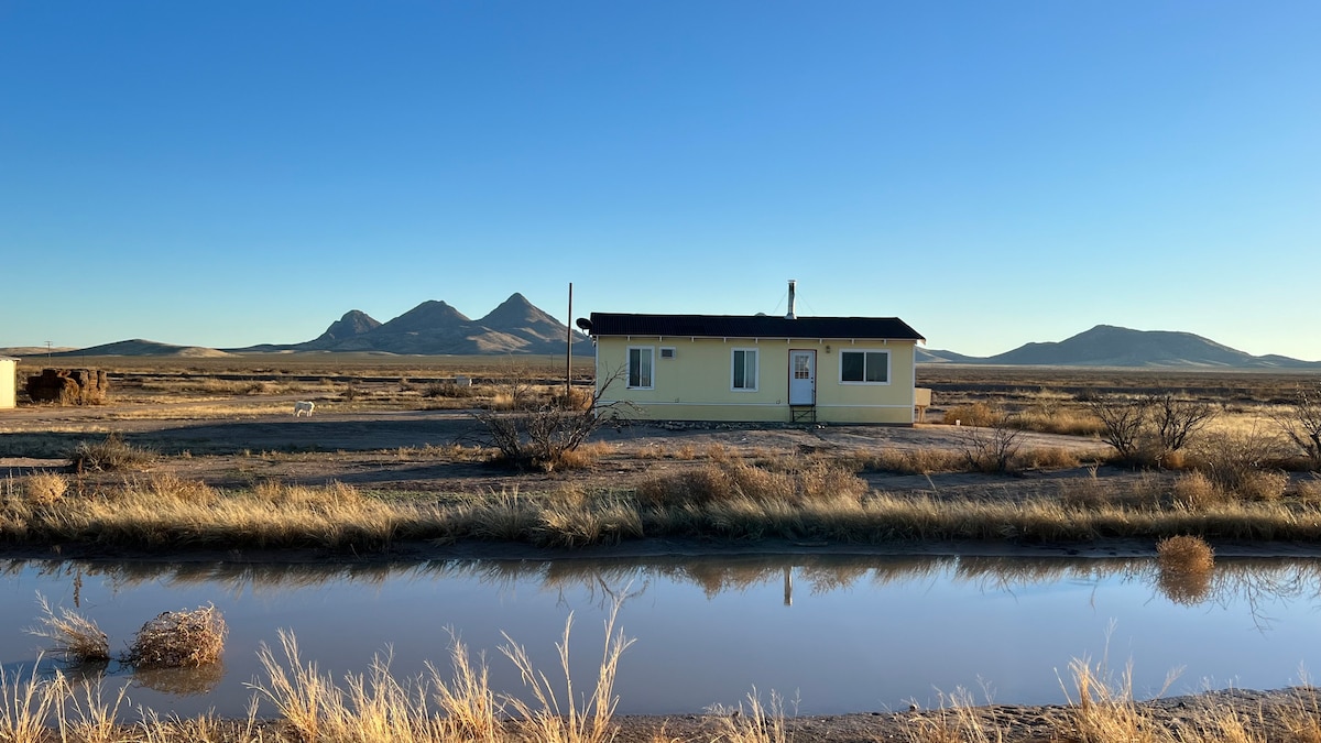 A rustic yellow cabin is set against a backdrop of rolling mountains under a clear blue sky. A small body of water reflects the cabin and landscape, while dry grass and sparse vegetation surround the area, emphasizing the tranquil, remote environment.