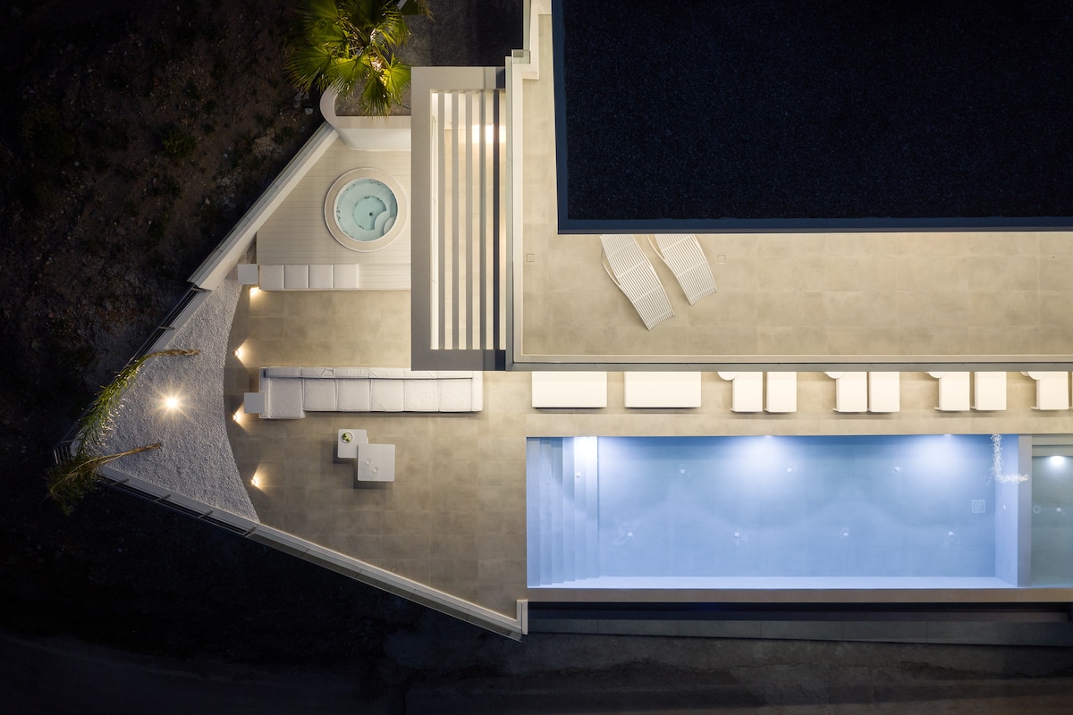 An aerial view of the outdoor living space showcases a heated spa and a spacious pool, bordered by loungers. The terrace features a stylish seating area, illuminated by soft lighting, emphasizing a relaxing atmosphere for enjoying evenings under the stars.