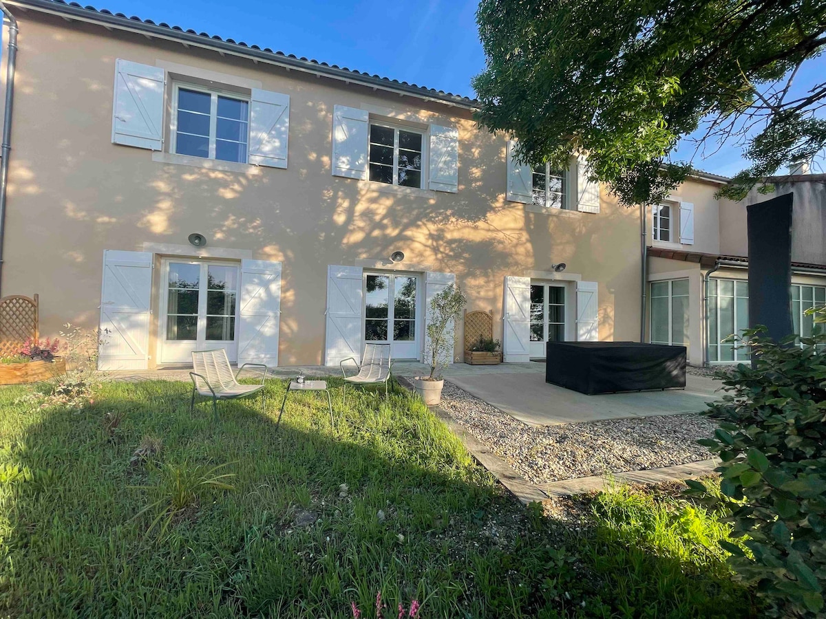 The exterior of the spacious two-story home features large windows framed by white shutters. A sunlit patio area is visible, surrounded by green grass and garden elements, equipped with lounge chairs. The tranquil setting is enhanced by nearby trees casting soft shadows.