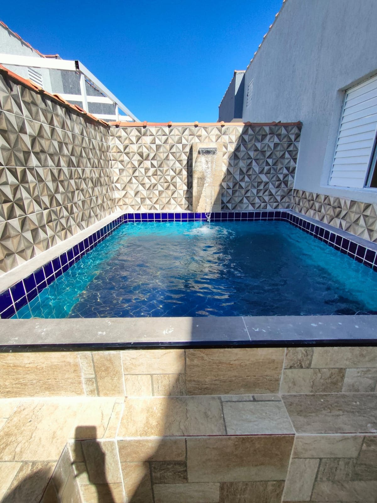 A private swimming pool is framed by textured tiled walls, featuring a calming water feature. The pool is surrounded by a blend of light and dark tiles, reflecting the clear blue sky above.