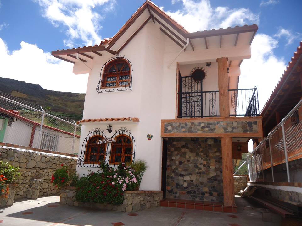 The exterior of a two-story cabin is shown, featuring a mix of stone and stucco finishes. Curved wooden windows and a decorative balcony adorn the upper level. Colorful flowers are planted along the entrance path, and a clear blue sky adds to the inviting atmosphere.