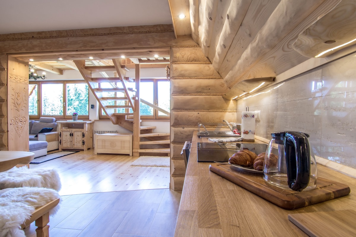 A modern kitchen area is highlighted by warm wooden finishes and natural light streaming through large windows. A wooden countertop displays a kettle and croissants. The space features decorative wooden accents, with a staircase leading to the upper level visible in the background.