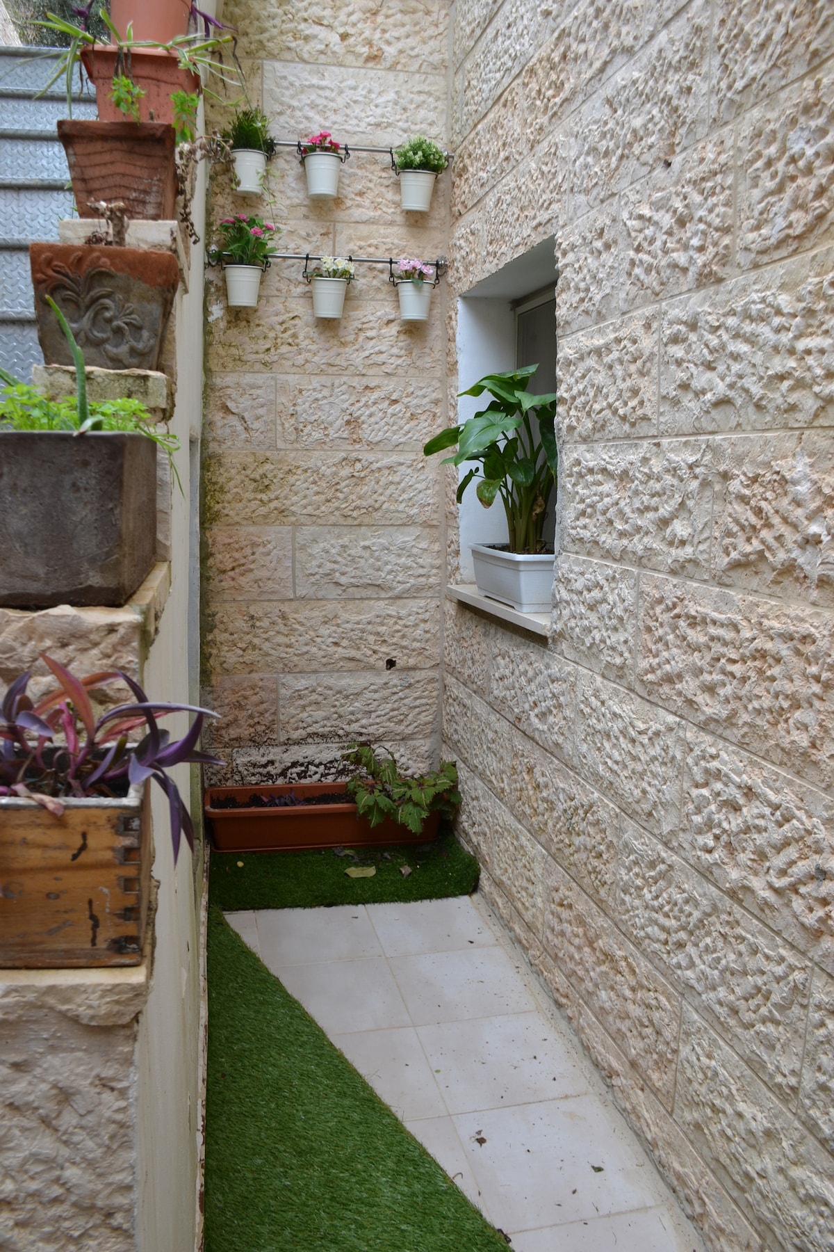 A narrow exterior corridor is framed by textured stone walls, adorned with planters featuring vibrant flowers. The pathway is lined with artificial grass, leading to a small window that showcases additional greenery. Natural sunlight brightens the space, enhancing the peaceful ambiance.