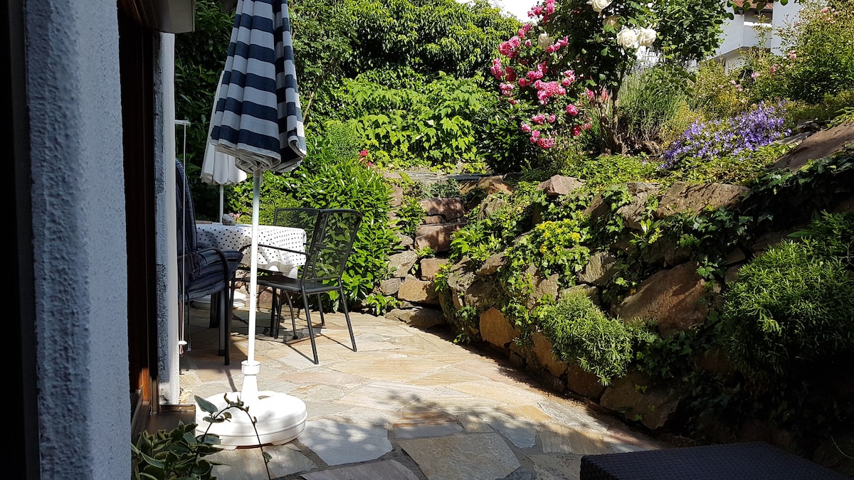 A private terrace is visible, surrounded by lush greenery and colorful flowers. Stone pathways lead through the garden, connecting the seating area with a table and chairs, and a large umbrella provides shade. Natural light enhances the inviting atmosphere of the outdoor space.