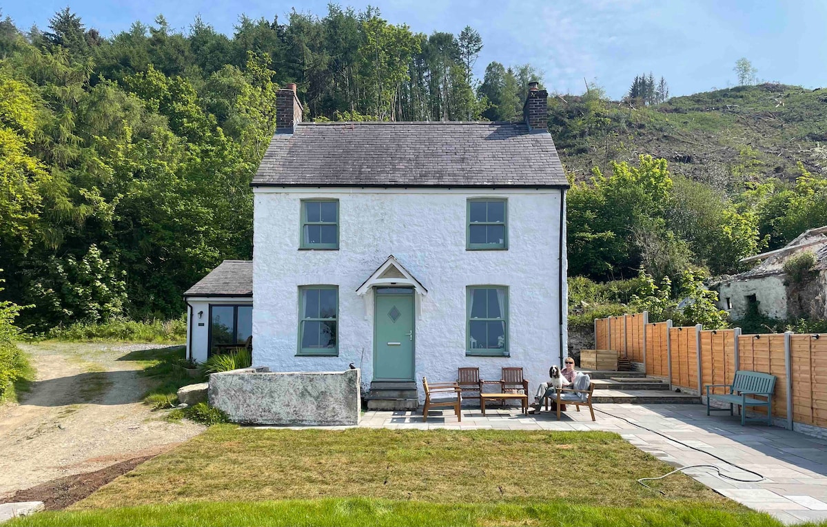 A modernised stone farmhouse is set amidst lush greenery, with a well-kept lawn in front. The exterior features light-colored walls and a welcoming front door, complemented by outdoor seating arrangements on a paved area. Natural light brightens the surrounding space.