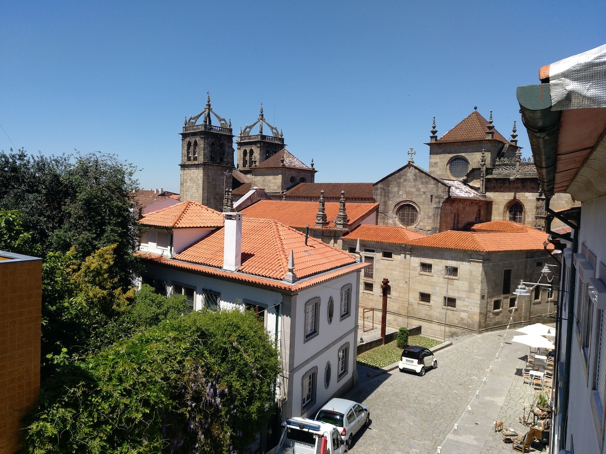 The image captures a view of Braga's Sé Cathedral, featuring its prominent towers topped with spires. A mix of terracotta roofs and stone buildings is visible, along with greenery and a narrow street that adds to the charm of the historic area.