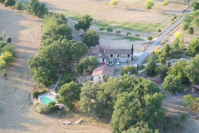 An aerial view of the property reveals a rustic stone building surrounded by trees and open fields. A tranquil pool is located nearby, with pathways leading to the home. The landscape features a mix of greenery and golden brush, creating a serene rural setting.