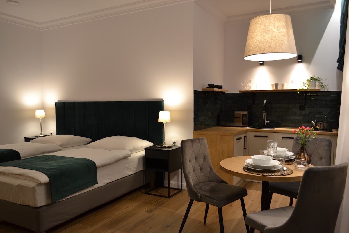 A studio apartment features a spacious bed with green accents and bedside lamps. A small dining table with two chairs is set, and a kitchenette is visible in the background, equipped with modern appliances. Soft lighting creates a cozy environment.