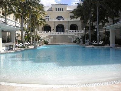 A large swimming pool reflects the surrounding architecture, framed by palm trees and lounge chairs. Steps lead up to a grand building with an expansive veranda, offering a glimpse of the elegant outdoor space designed for relaxation.