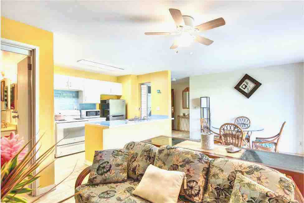 A bright living area features a floral-patterned sofa and a ceiling fan overhead. Open cabinetry reveals a modern kitchen in the background. Natural light fills the space, highlighting the cheerful yellow walls and a round dining table with wicker chairs along with a decorative piece on the wall.