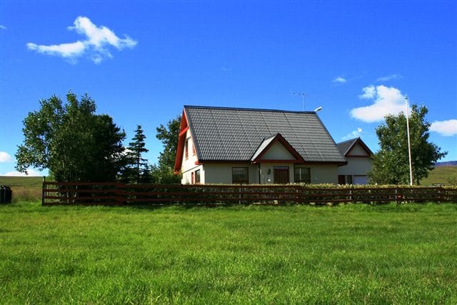 A two-story house is surrounded by a lush green lawn, featuring trees on either side. The building exhibits a gabled roof with red accents and a white exterior, complemented by a wooden fence that outlines the property. Clear blue skies provide a vibrant backdrop.