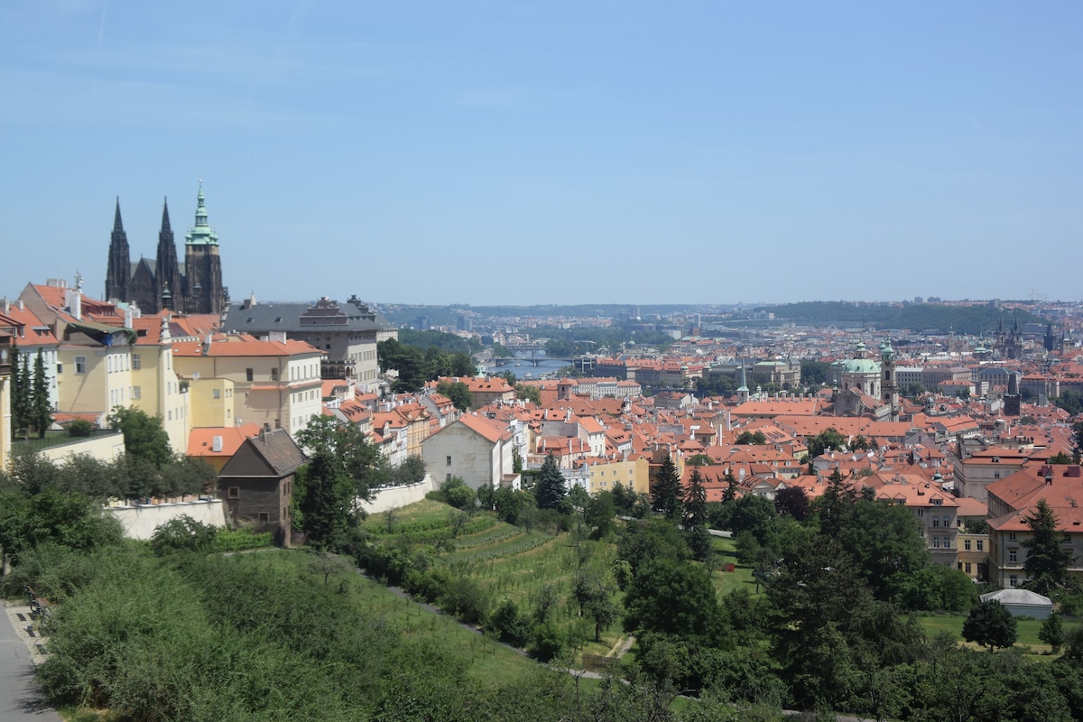The panoramic view captures a vibrant cityscape of Prague, showcasing terracotta rooftops and green spaces. The iconic spires of Prague Castle rise in the background, accompanied by lush hills and scattered residential buildings, indicative of the city's historic architecture.