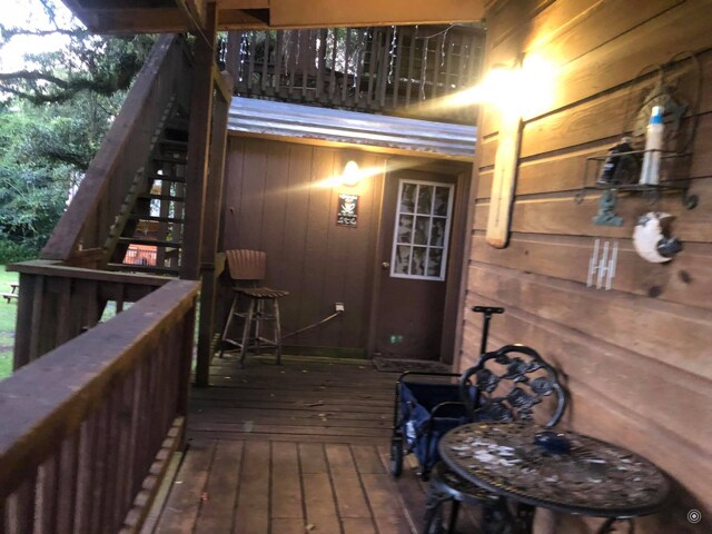 A rustic exterior showcases wooden paneling with a warm light illuminating the entryway. A small table and chairs are placed on the porch, while a staircase leads to the upper level. Near the door, a decorative wall piece and bar stool are visible.