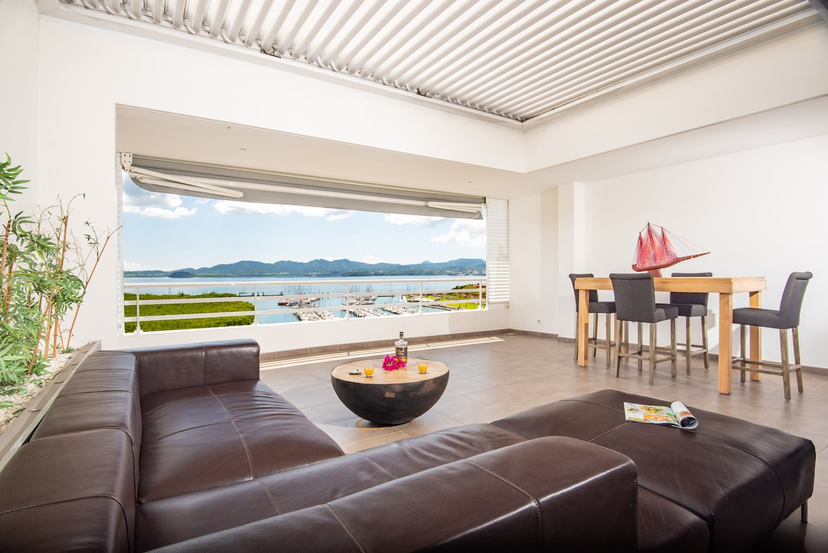 A spacious living area is highlighted by panoramic views of the Caribbean Sea, visible through large windows. A comfortable sectional sofa faces a round coffee table, while a dining table for six is positioned nearby, creating a welcoming space for relaxation and meals.