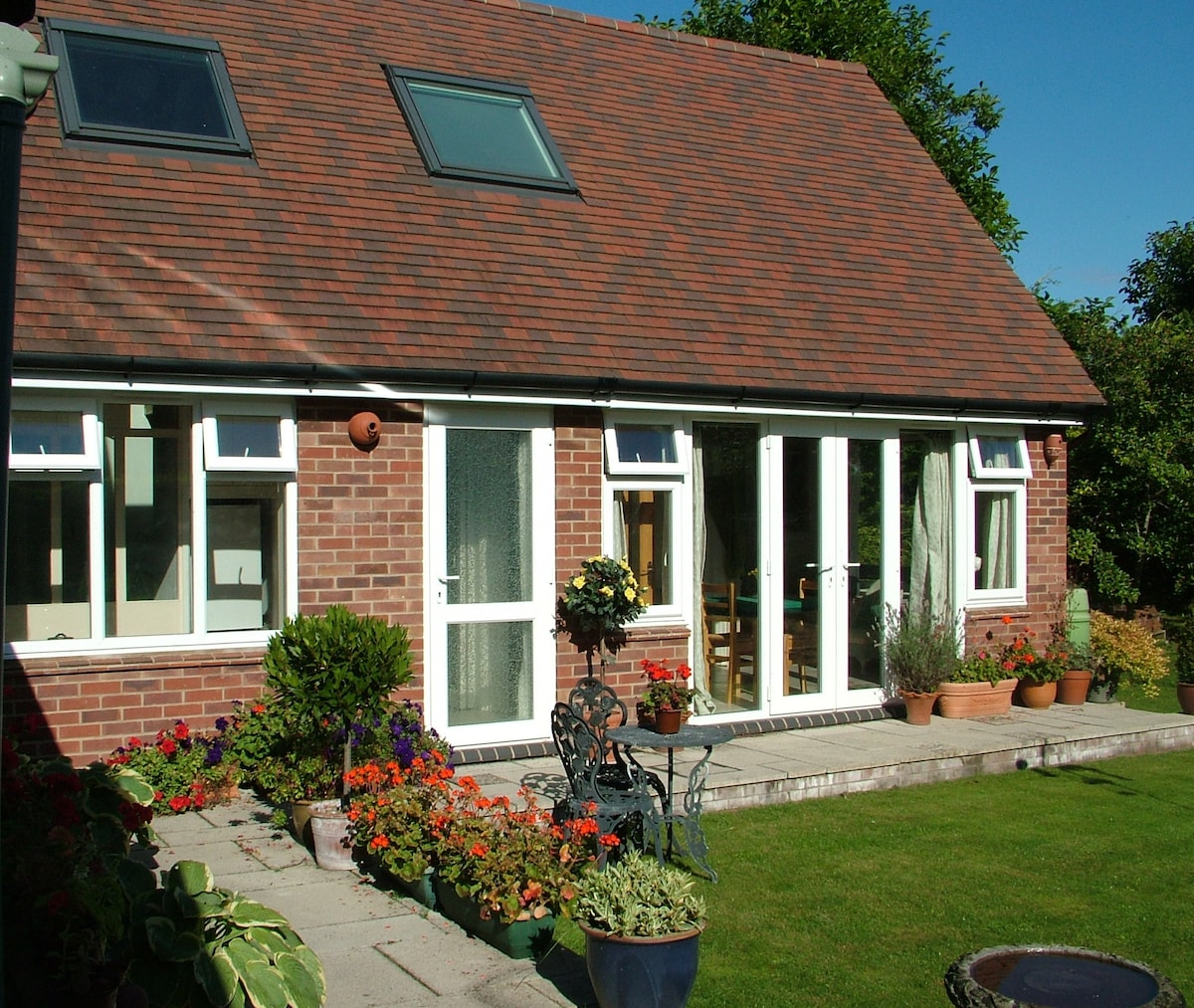 A charming exterior of The Lodge is shown, featuring a well-maintained garden with colorful flowers and potted plants. Large glass doors open to a bright interior, complemented by windows beneath a sloped roof. A seating area with chairs is visible on the patio.