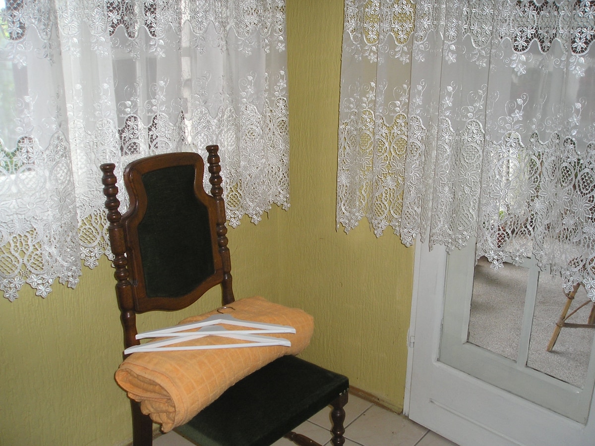 A wooden chair with a dark seat cushion is positioned near a lace curtain, allowing soft light to filter through. A neatly folded orange towel rests on the chair, while a glass door opens to a patio area, enhancing natural light in the space.
