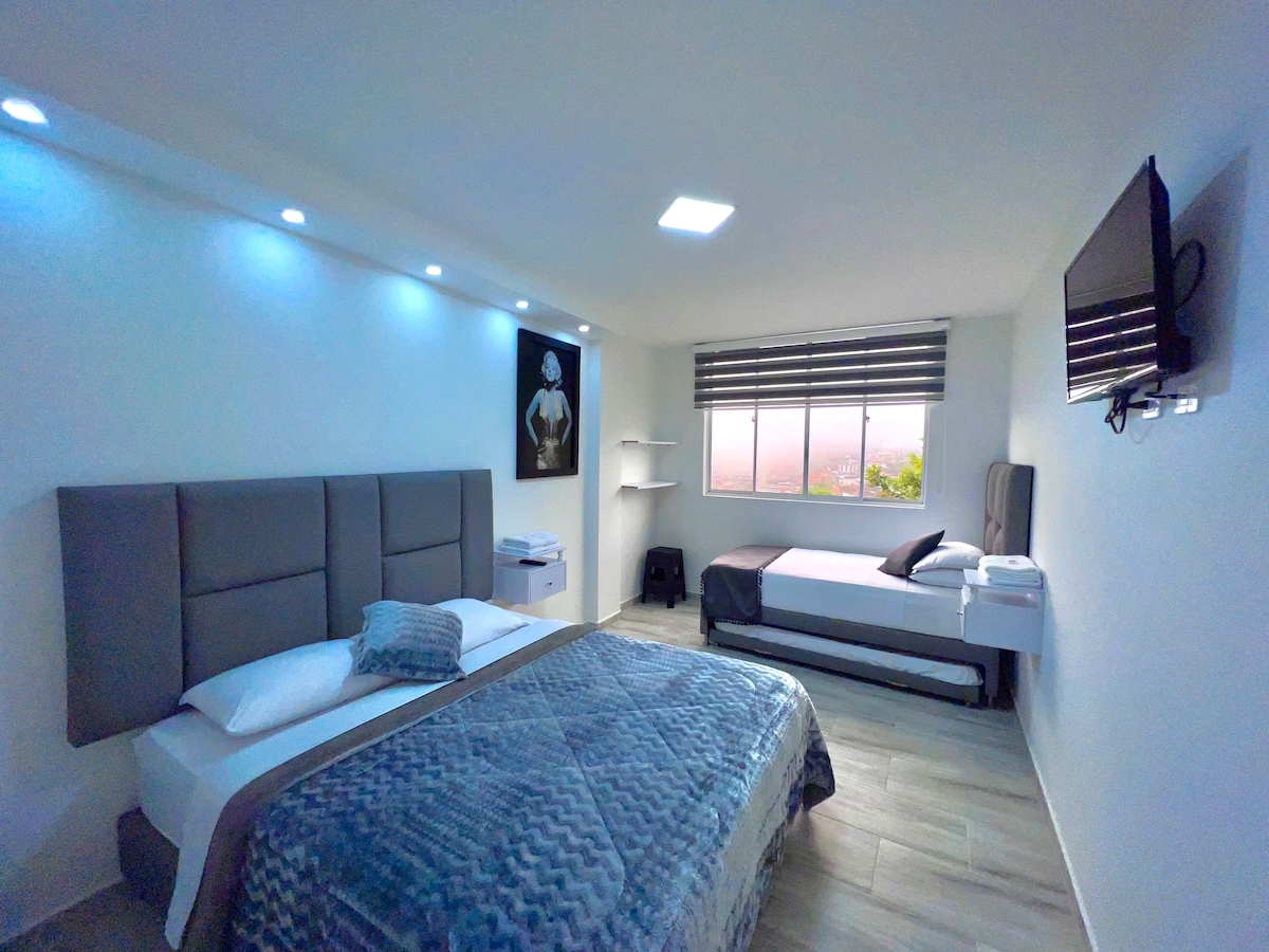 A well-appointed sleeping area features a spacious bed with a textured blanket and a single bed nearby. Wall-mounted lighting creates a soft glow, while a large window offers a view. A television is mounted above a minimalist white shelf, enhancing the room's functionality.