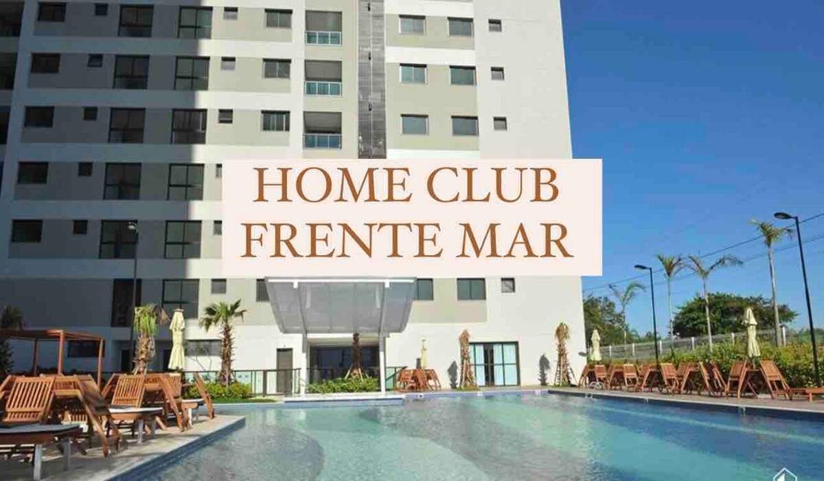 A modern building is showcased, featuring large windows and a glass entrance. A spacious pool is visible in the foreground, surrounded by lounge chairs. Tropical plants line the area, adding greenery to the resort-style ambiance. The location is in front of the beach.