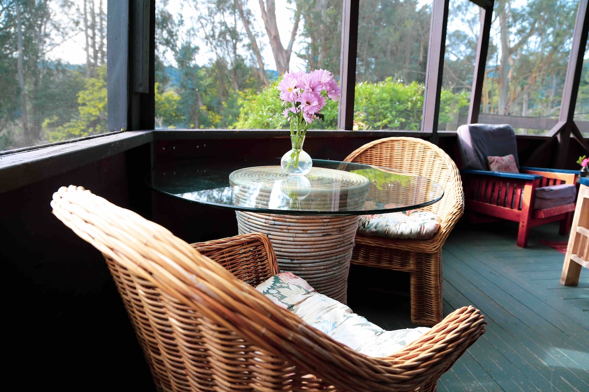 A screened porch features a round glass table surrounded by two wicker chairs, each with cushioned seating. A small vase with fresh flowers is placed at the center of the table, offering a view of the surrounding greenery through the screens.