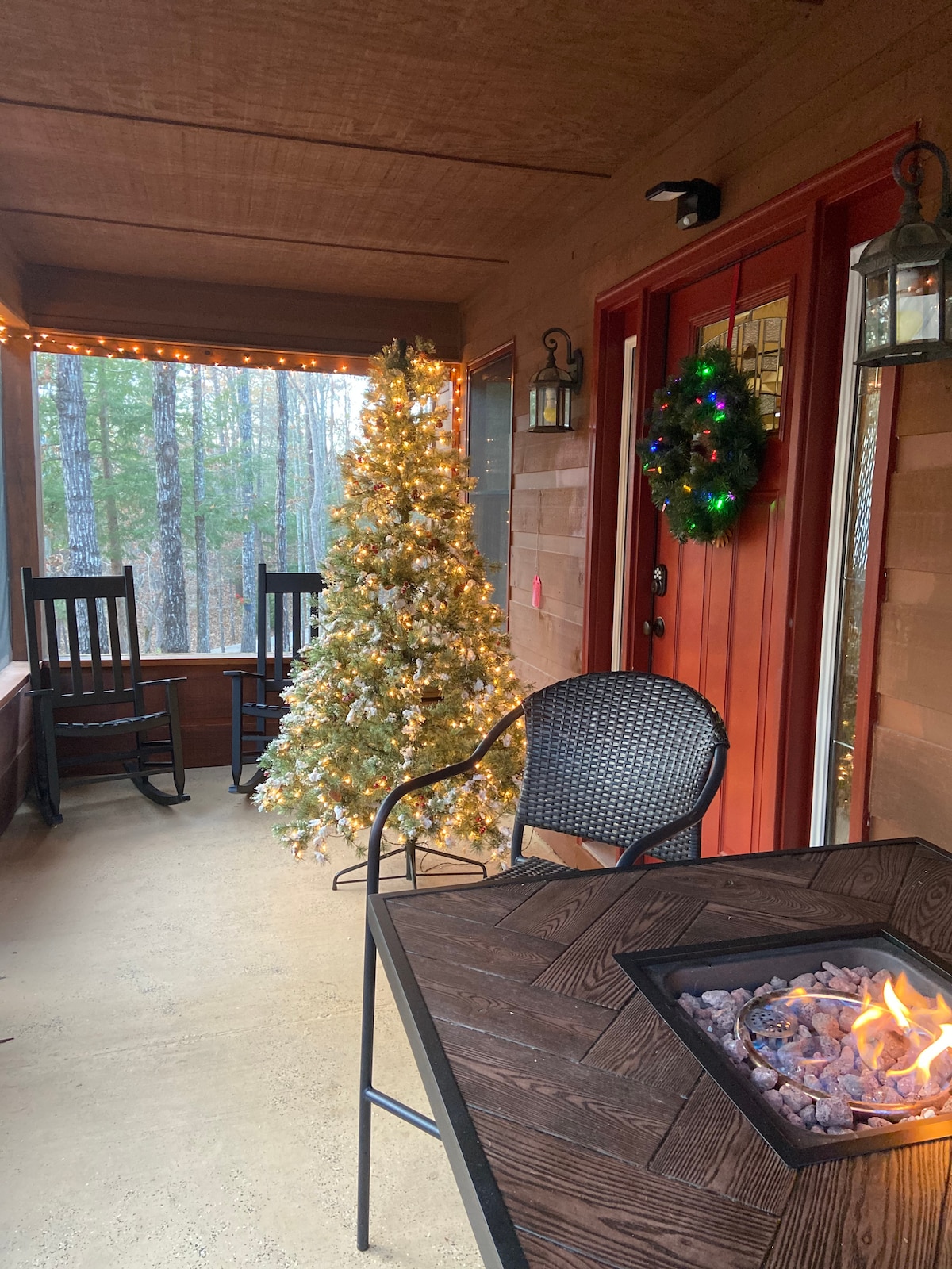 A screened-in porch features two rocking chairs beside a table with a gas fire pit. A decorated Christmas tree stands nearby, adorned with twinkling lights. A green wreath hangs on the door, complementing the warm, inviting atmosphere of the outdoor space.